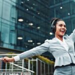 Frau in Business-Outfit mit Kopfhoerern streckt die Arme aus und lacht vor einem modernen Buerogebaeude – vital durch Bewegung mit personal trainer koeln